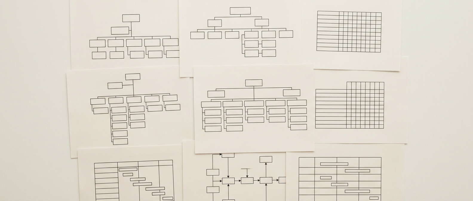 Printed technical documents overlapping in a loose grid, seen square-on from above: organisation charts of empty boxes, process diagrams and blank matrices, in flat shadowless light.